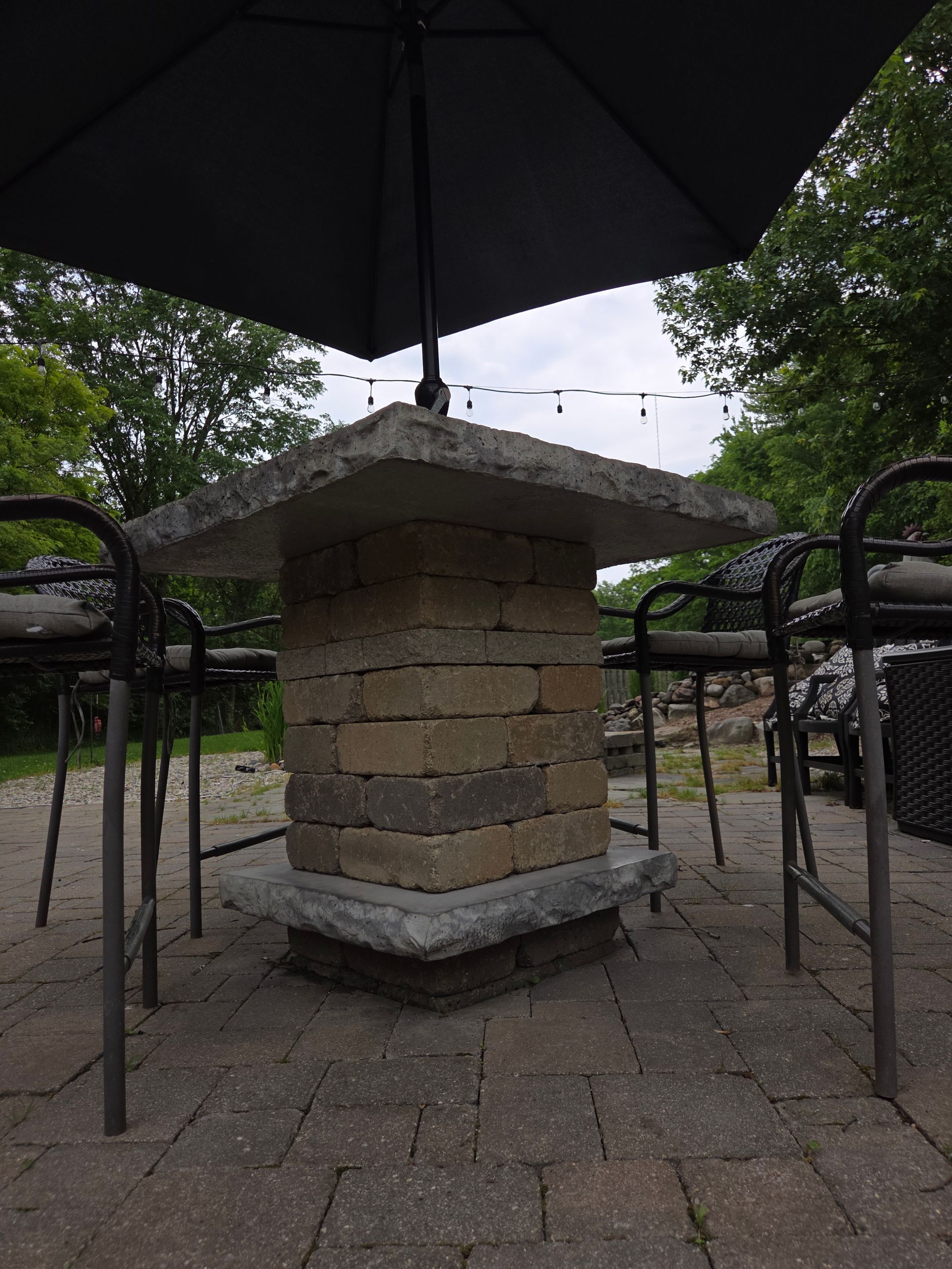 Stone pedestal table under a black umbrella in an outdoor park setting, with metal chairs around it.