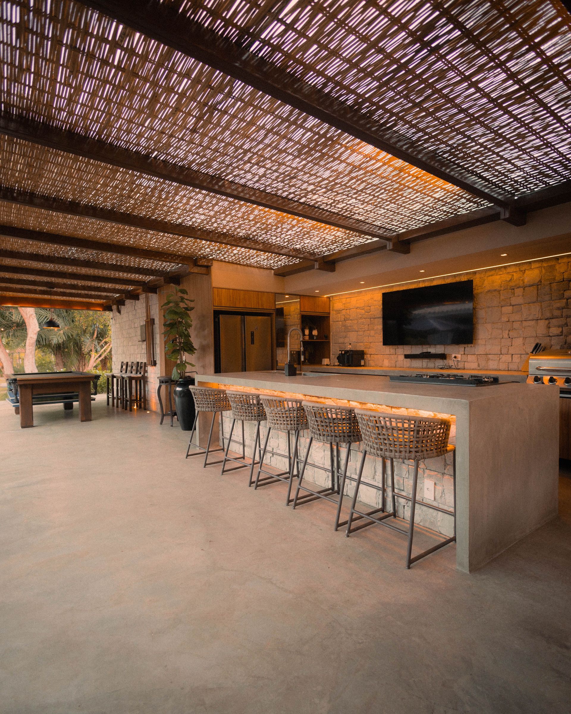 Open-air rustic patio bar with woven roof, stone counter, metal stools, and lounge seating