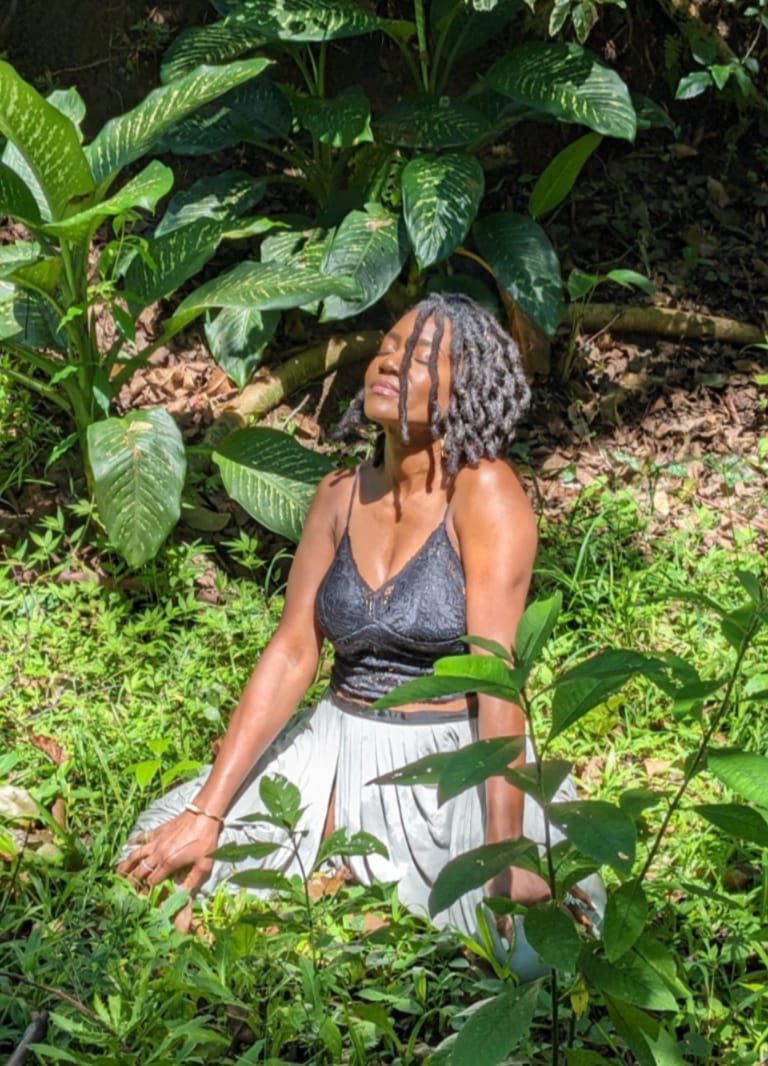 Amber Wood sitting in lush greenery, wearing a black top and light skirt, looking upward in sunlight