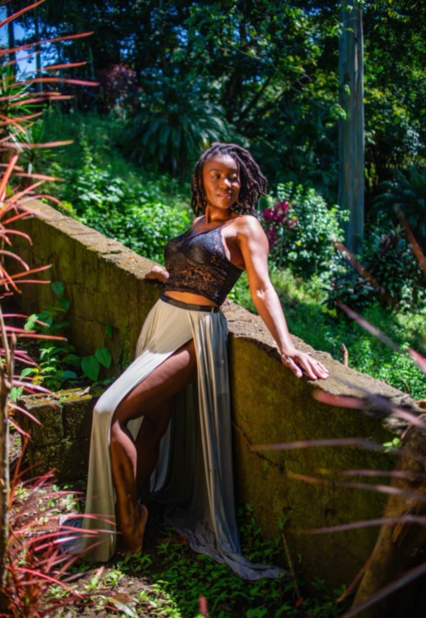 Amber Wood posing on a stone wall in a lush garden, wearing a black top and flowing beige skirt.