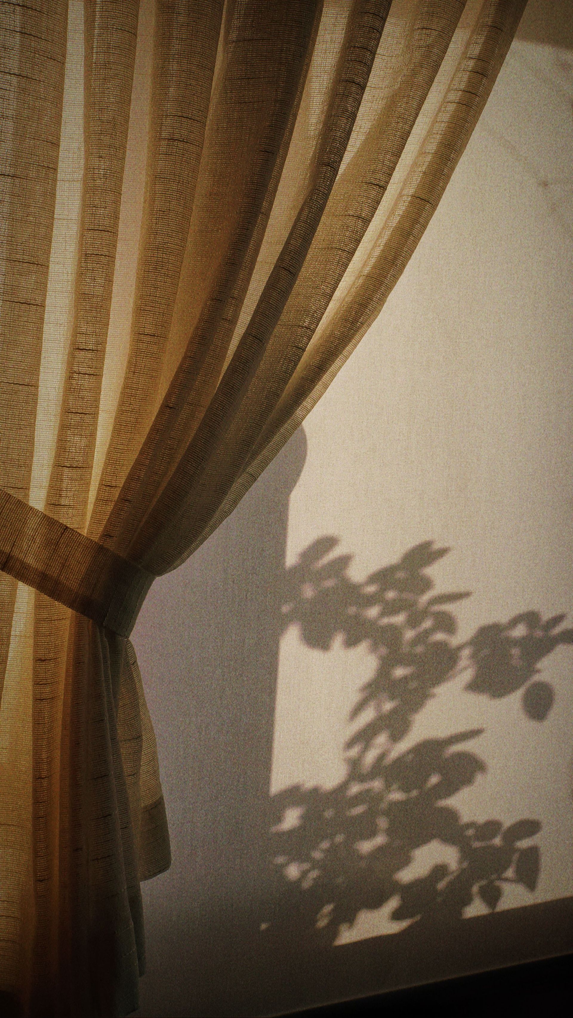 sheer beige curtain drawn to once side to reveal light shining through a window casting a shadow