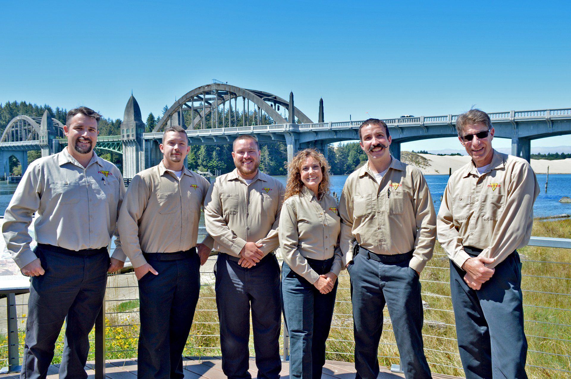 Group of six people in tan shirts and dark pants posing with a bridge in the background.