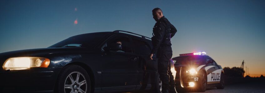A police officer stands by the driver's side door of a dark car at dusk, with a patrol car’s lights flashing behind them.