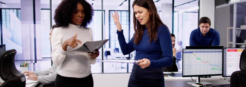 Two colleagues in a modern office discuss documents, one appearing frustrated while gesturing with her hand.