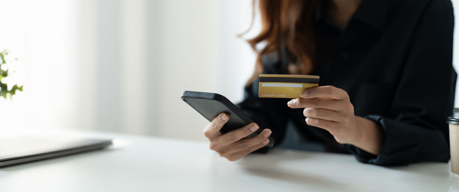 A person in a black shirt holds a credit card in one hand and a smartphone in the other, likely making an online purchase.