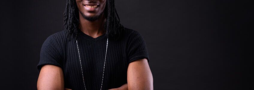 A smiling person with dreadlocks wearing a black v-neck t-shirt and a long chain, arms crossed against a black background.