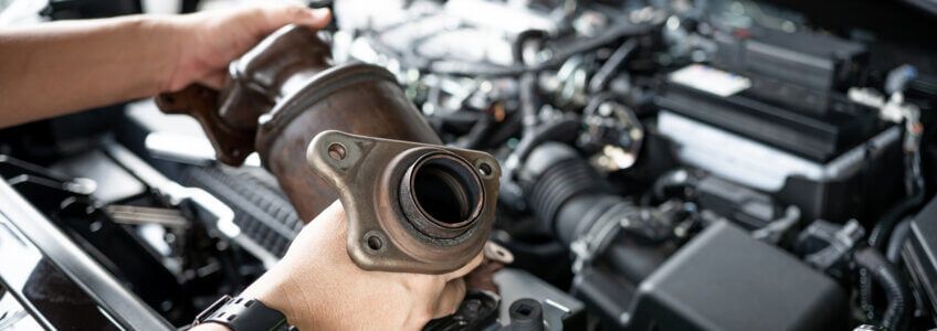 A mechanic holds a removed catalytic converter over an open car engine bay.