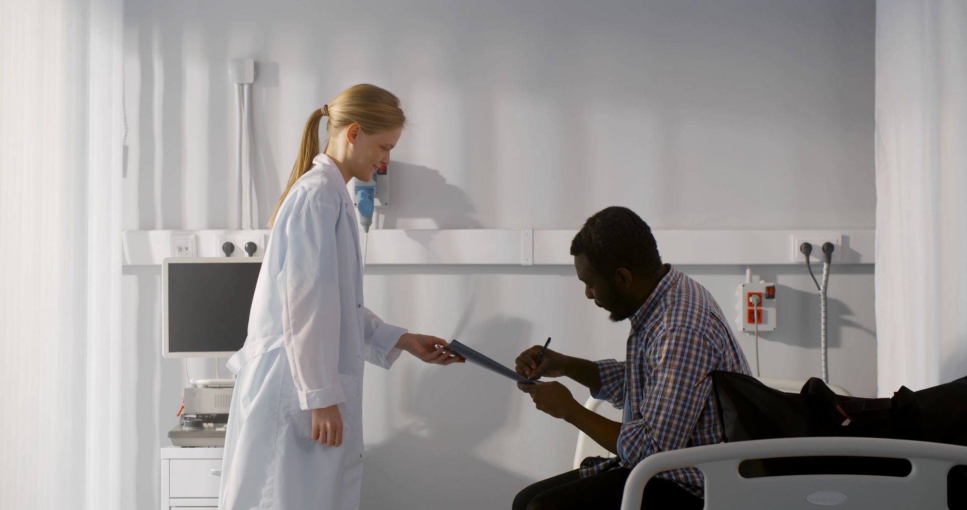 doctor in a white coat hands a clipboard to a person sitting on a hospital bed, who is signing a document to be disharged