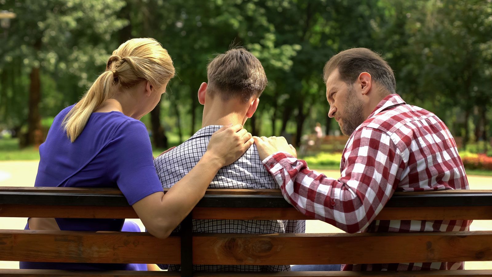 Two adults sit on a park bench, each with a hand on the shoulders of a person sitting between them in a supportive gesture.