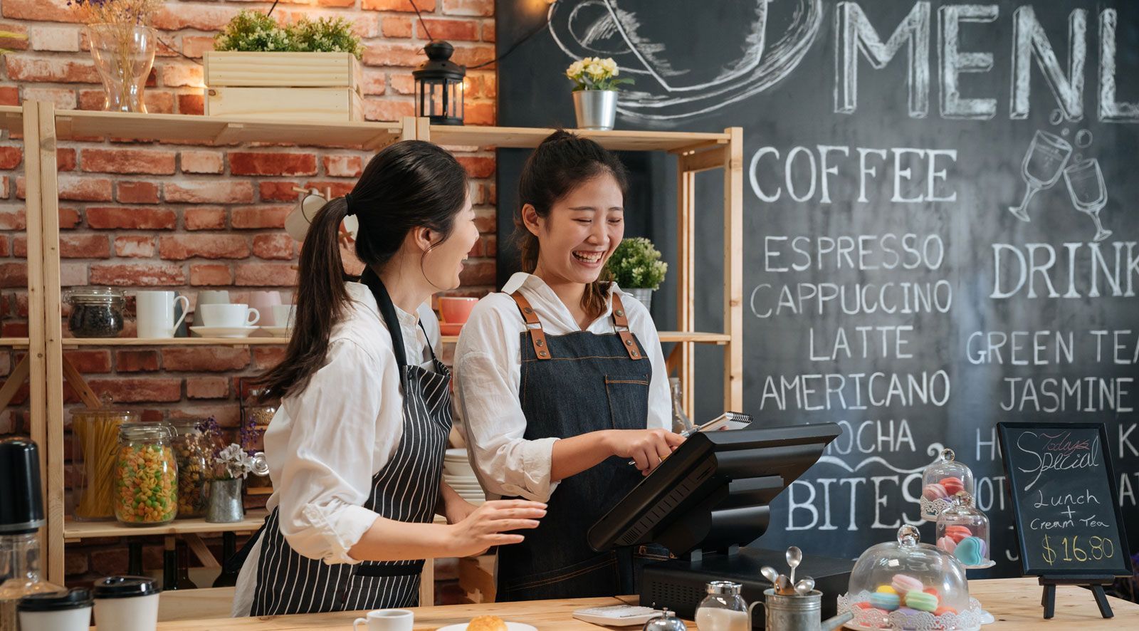 Two baristas in aprons laughing while using a point-of-sale system in a coffee shop with a chalkboard menu.
