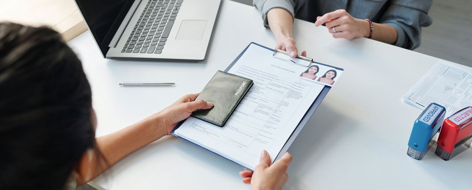 A person hands a document and a small black wallet to another person across a desk with a laptop and two rubber stamps.