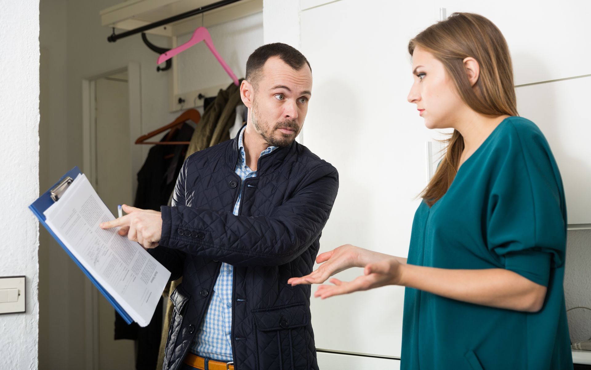 A person points to a document on a clipboard while speaking to someone in a home entryway.