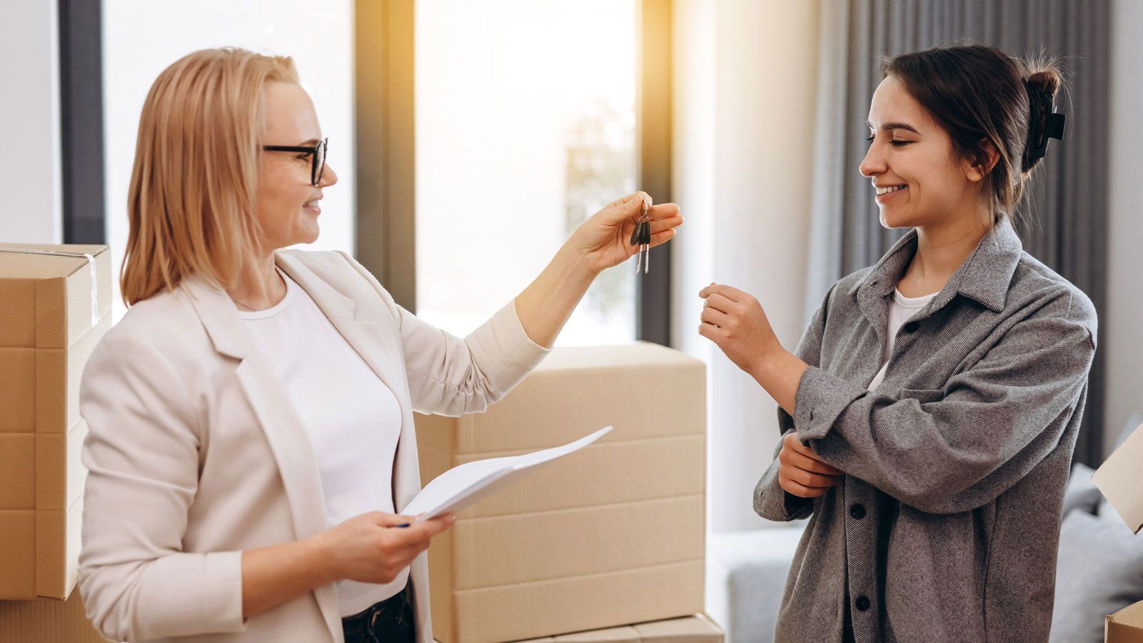 A person hands a house key to another person in a room filled with cardboard moving boxes.