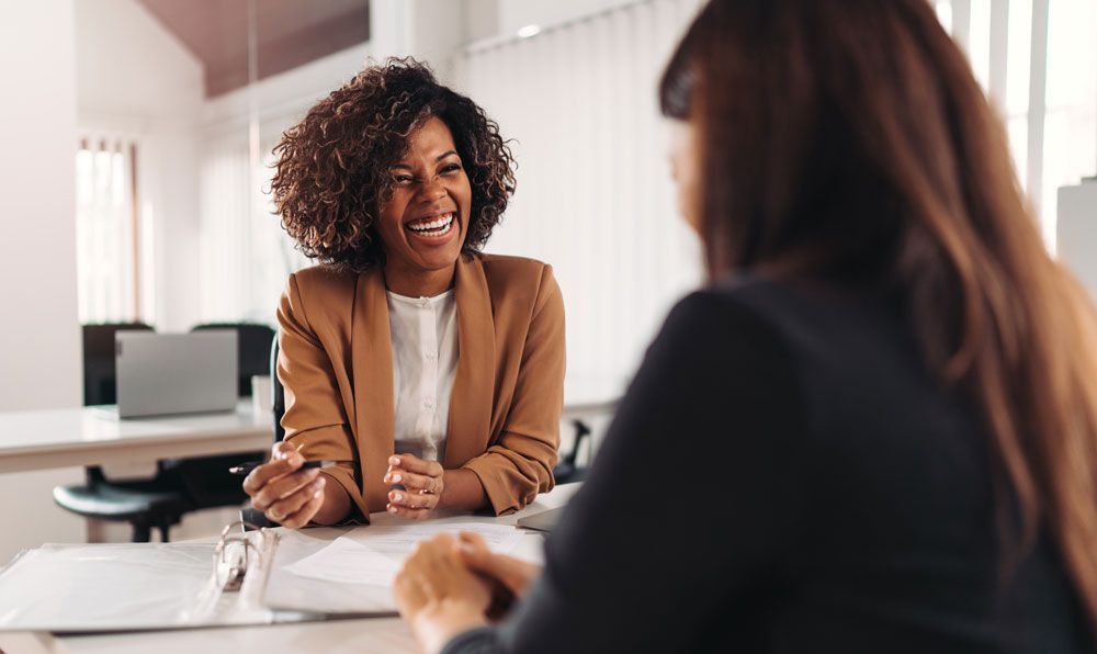 Two colleagues in business attire smile while sitting at a desk and collaborating in a bright, modern office.