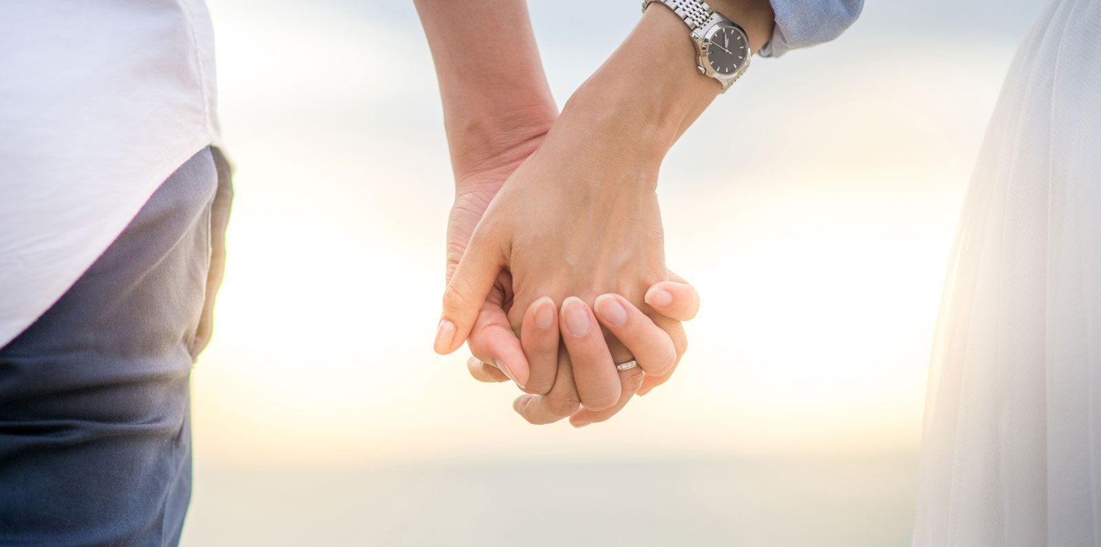 A pair of hands held together against a soft, sunlit background, with one person wearing a watch and a ring.
