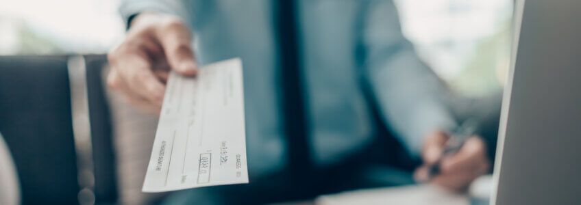 A person in a professional shirt holds out a personal check toward the viewer while holding a pen in their other hand.