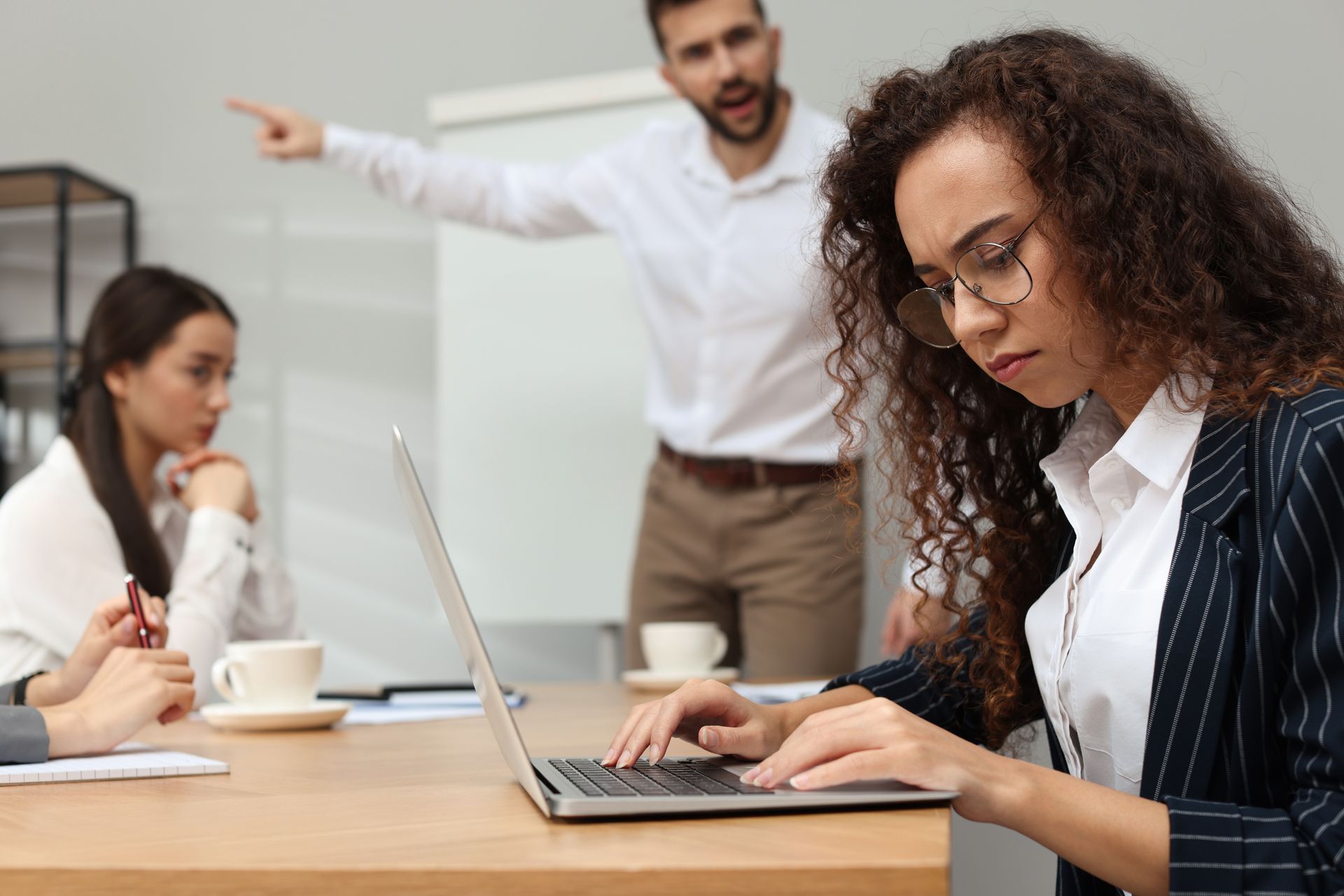 A man points across a meeting room at a woman typing on a laptop, while another person sits quietly nearby.