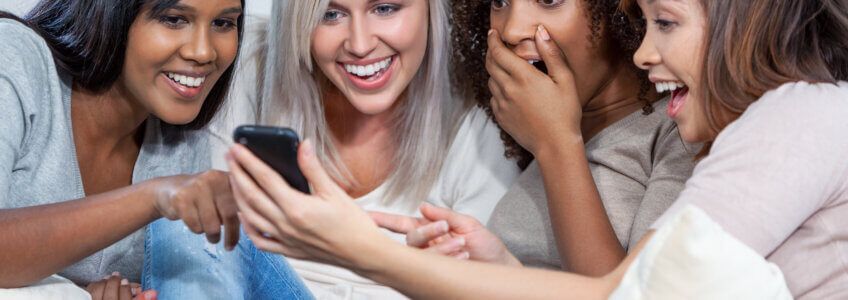 Four people huddled together, smiling and looking with excitement at a smartphone screen.