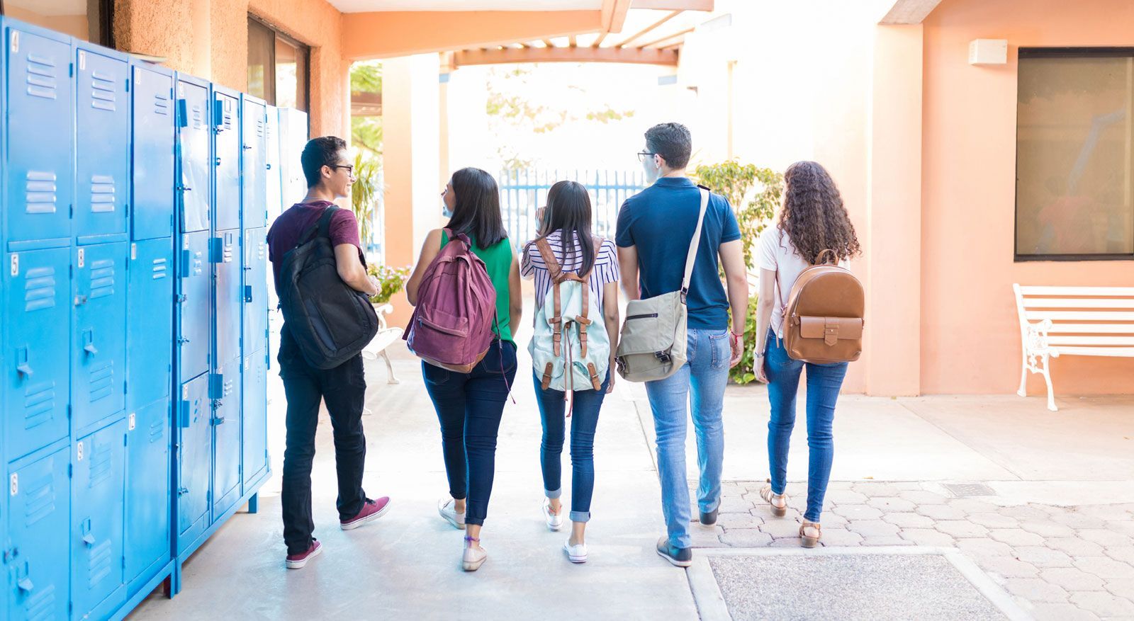 Five students walk away from the camera down a hallway lined with blue lockers, carrying backpacks.