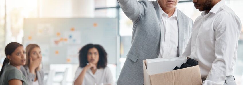 A person carries a box of belongings away as a superior gestures toward the exit in an office setting.