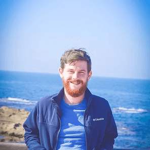 Smiling person in blue jacket standing by the ocean on a sunny day