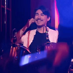 Smiling drummer onstage under purple and pink lights, holding a hand to his ear while seated at a drum kit