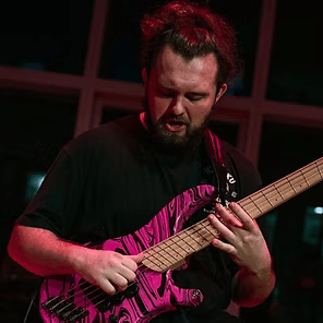 Musician playing a pink zebra-striped electric bass on a dark stage, lit by red light.