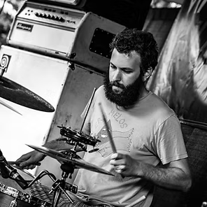 Man drumming in a rehearsal space with amps and a drum kit, shown in black and white.