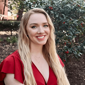 Smiling blonde woman in a red dress standing outdoors near greenery