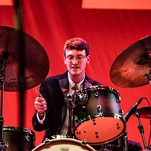 Drummer in a dark suit plays behind a drum kit on a red-lit stage