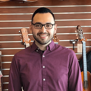 Smiling person in glasses and a purple shirt standing indoors with guitars on the wall