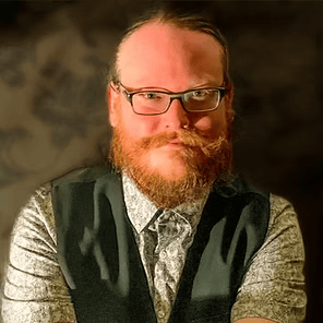 Portrait of a bearded man with glasses, wearing a vest and patterned shirt against a warm brown background
