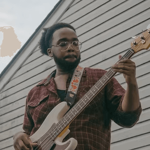 Person playing a white bass guitar outdoors beside a light gray building