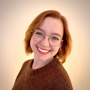 Smiling person with short red hair and glasses wearing a brown sweater against a beige background