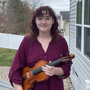 Person holding a violin outdoors beside a house, wearing a purple blouse and smiling.