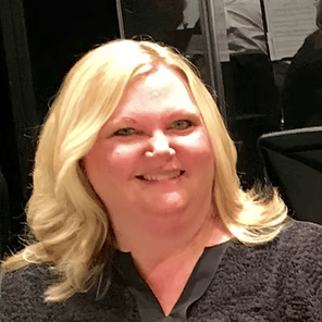 Smiling blonde person in a dark top, posing indoors against a dark background