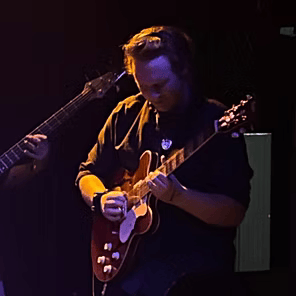 Musician playing a red electric guitar on a dark stage under purple lighting