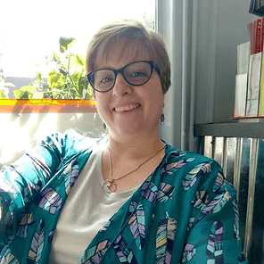 Smiling person in glasses and teal patterned top, sitting indoors by a sunlit window.