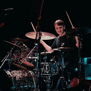 Smiling drummer on a dark stage, surrounded by a large drum kit and cymbals under spotlights