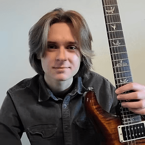 Person holding a sunburst electric guitar, seated indoors against a gray wall, looking at the camera.