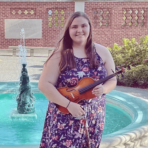 Woman holding a violin beside a fountain in a courtyard, wearing a floral dress and smiling.