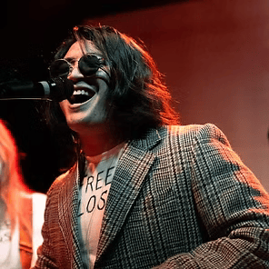 Singer in sunglasses and plaid blazer performs at a microphone under red stage lighting