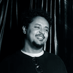 Smiling person in a dark sweater stands before black curtains.