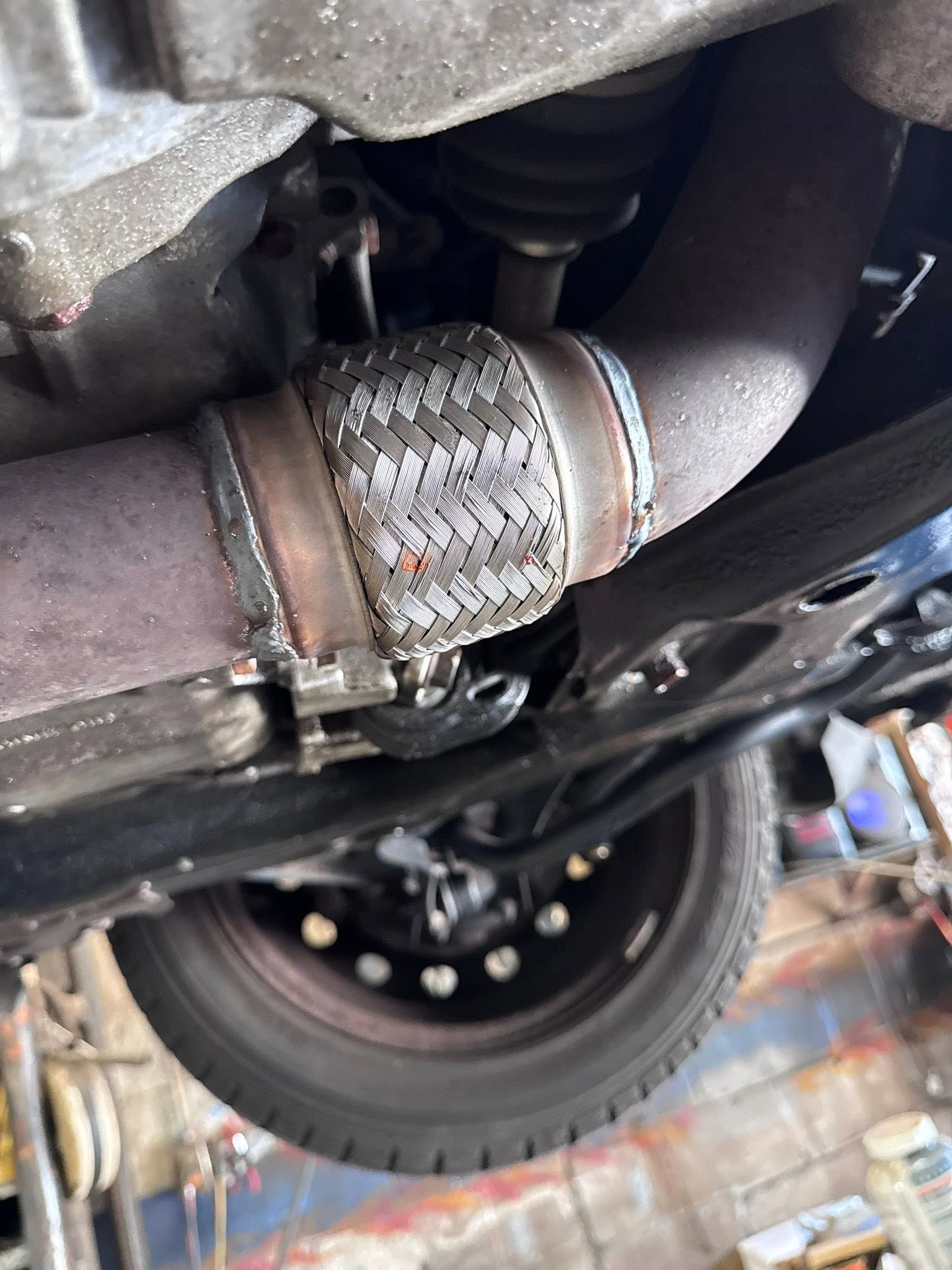 Close-up of a car’s exhaust pipe with a braided flex joint and the rear wheel visible underneath.