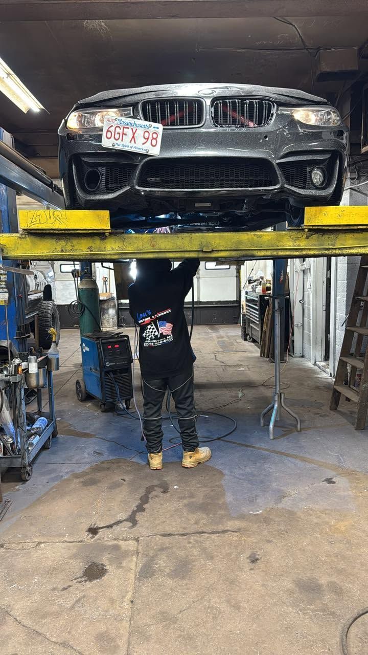 Black car raised on a lift in an auto shop, with a mechanic working underneath.
