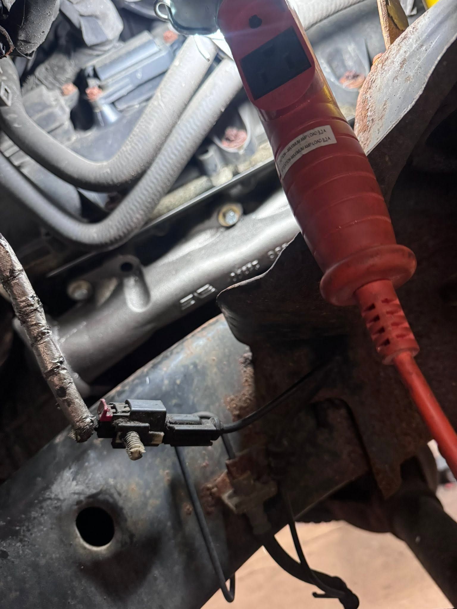 Red test probe touching a rusted metal engine bay with hoses and wiring cables.