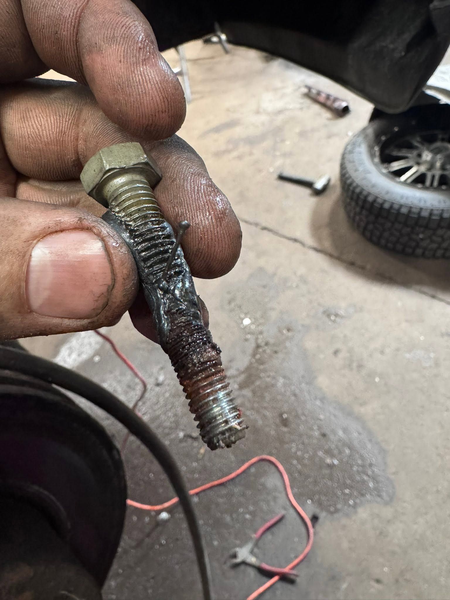 Hand holding a corroded, grease-covered bolt in a workshop floor area with tools and a wheel in the background