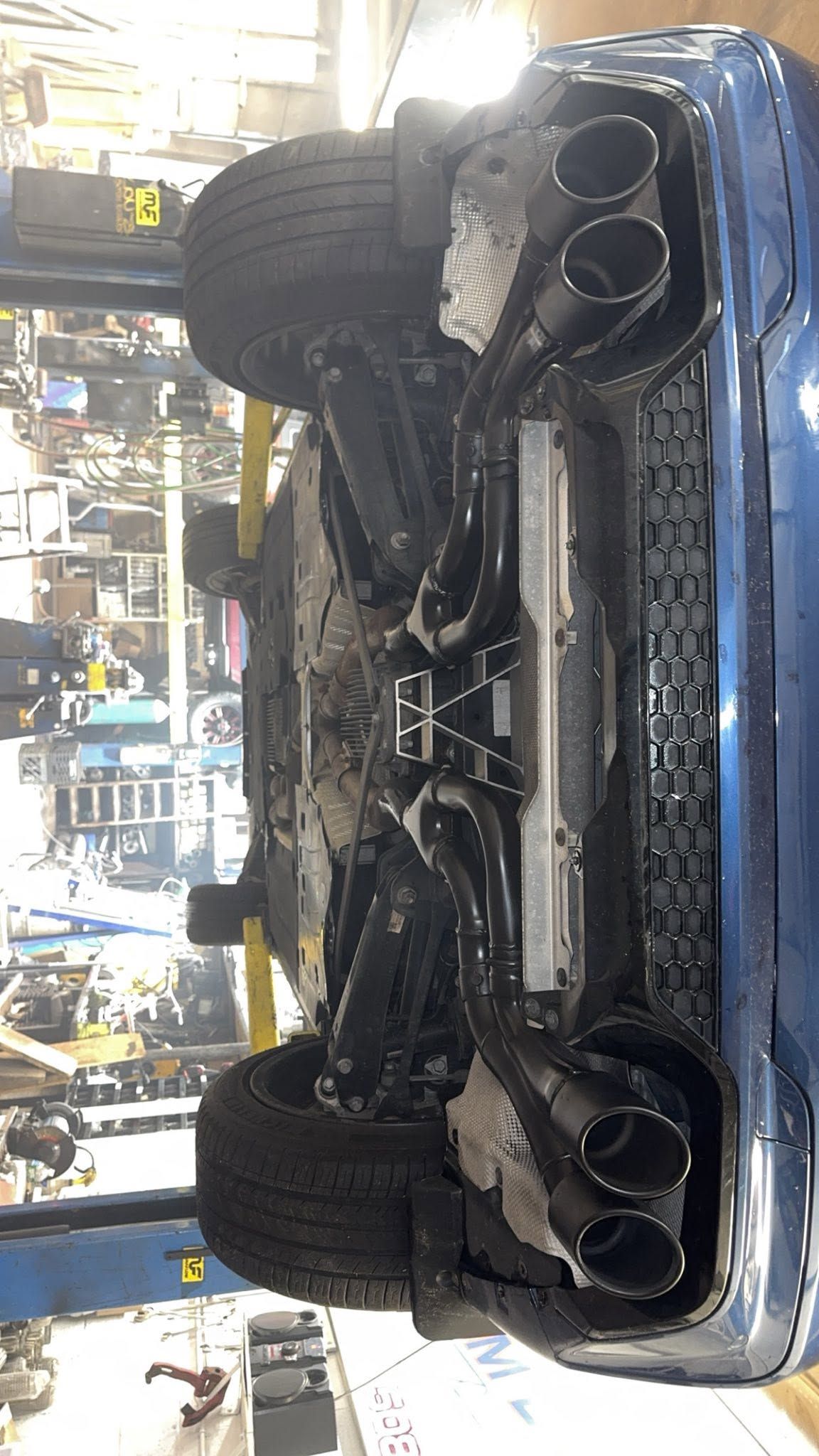 Undercarriage of a car on a lift in an auto shop, with twin exhaust tips visible and a spare tire mounted below.