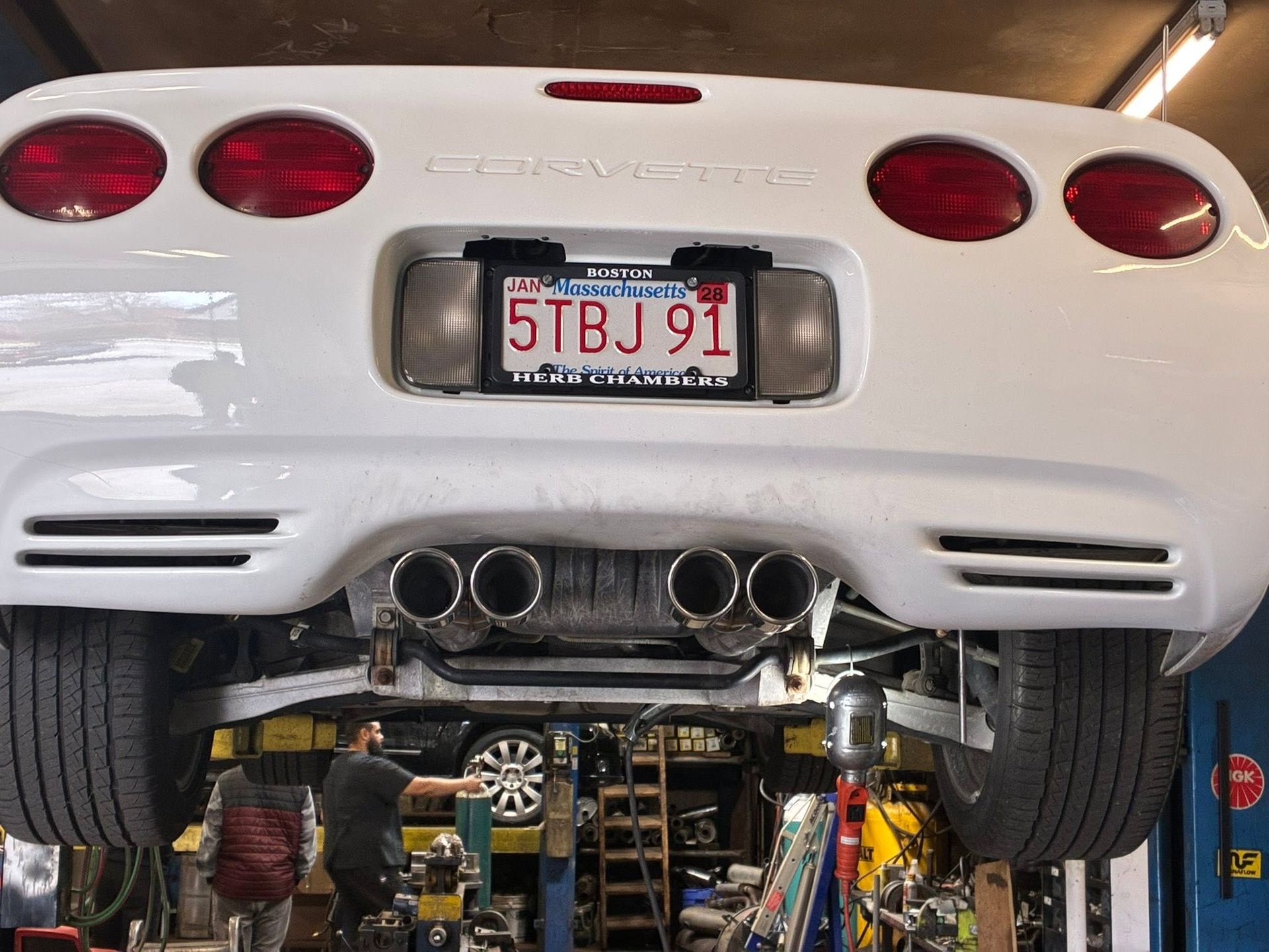 Rear view of a white sports car on a lift in a garage, showing dual exhaust and a Massachusetts plate.