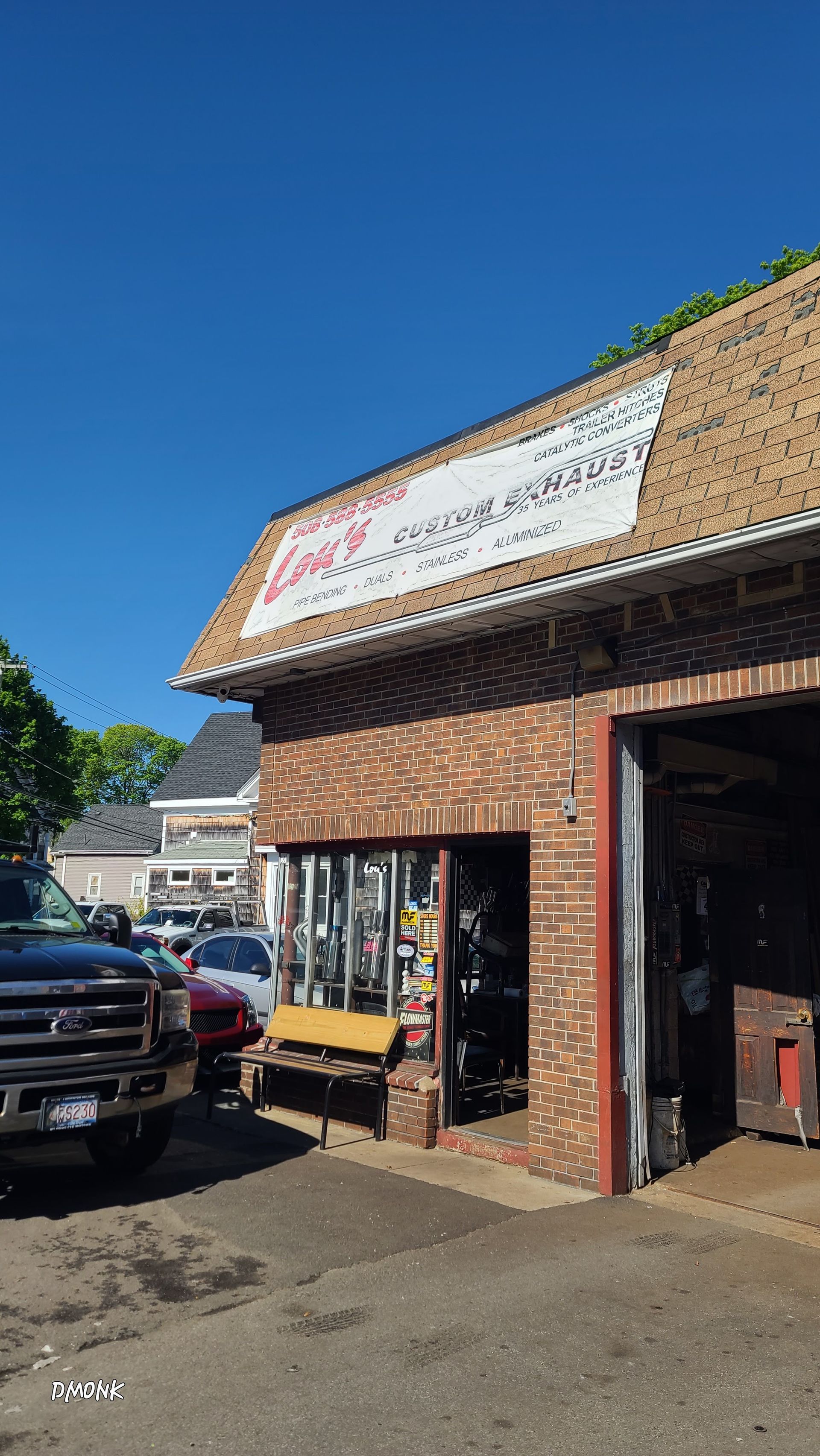 Brick storefront with a white sign, open garage-style entrance, and parked vehicles in bright sunlight